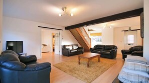 A spacious living room with a wooden floor, a black leather sofa, a wooden coffee table, and a staircase leading to an upper level.