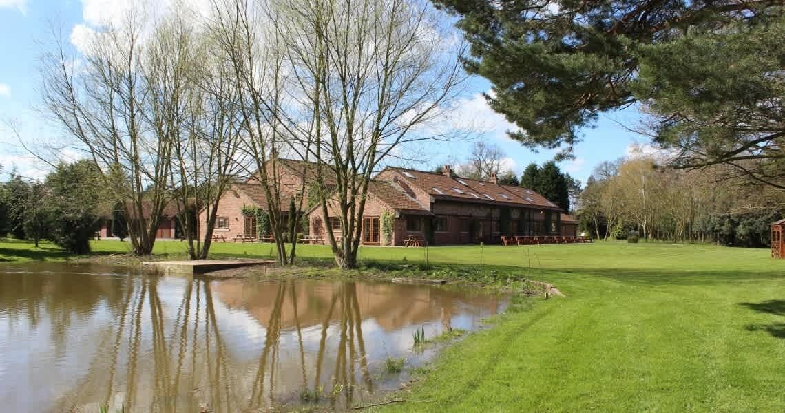 A building with a pond in front, surrounded by trees and a grassy area.
