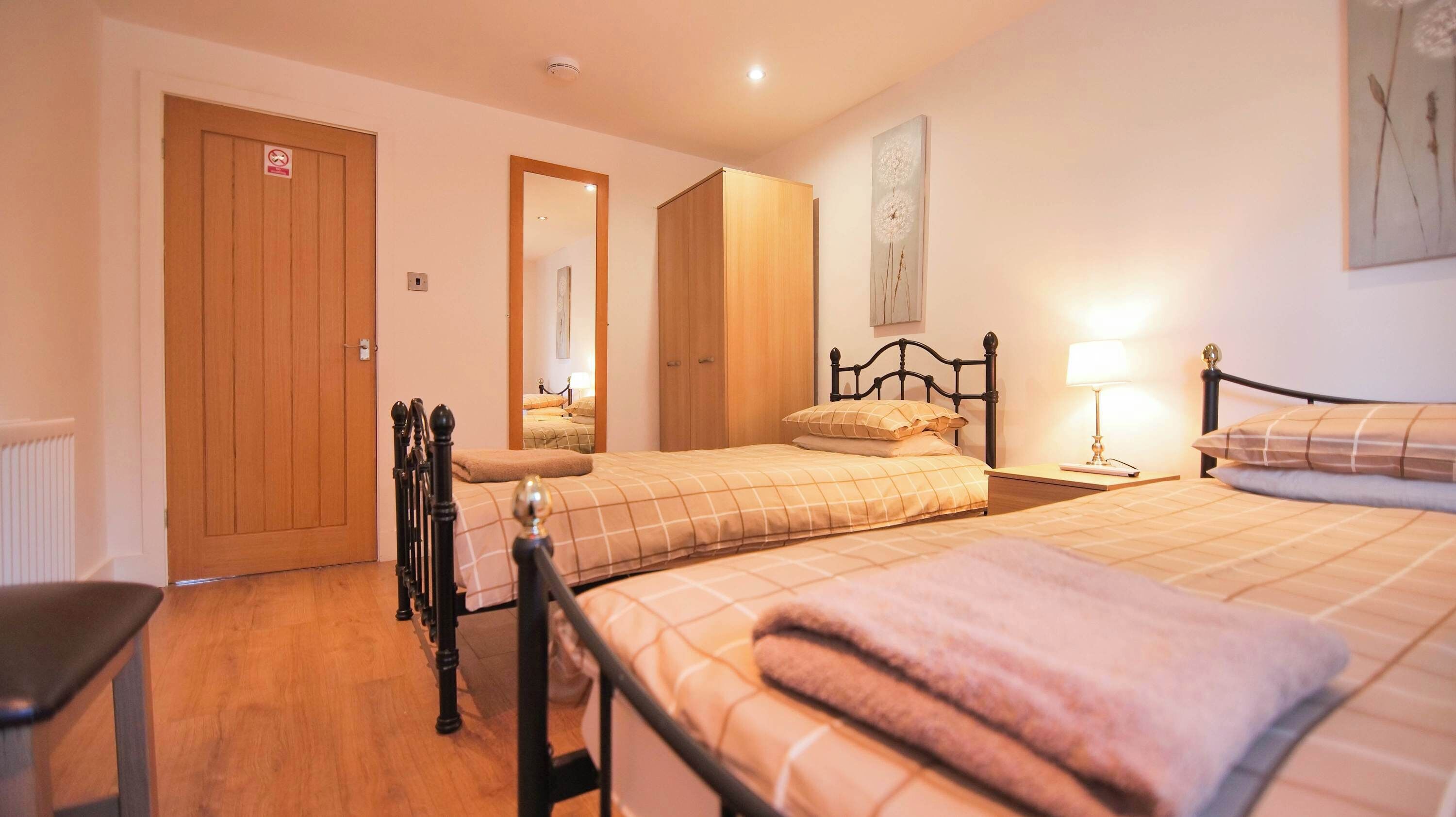 A bedroom with two beds, a wooden wardrobe, a mirror, and a bedside table with a lamp.