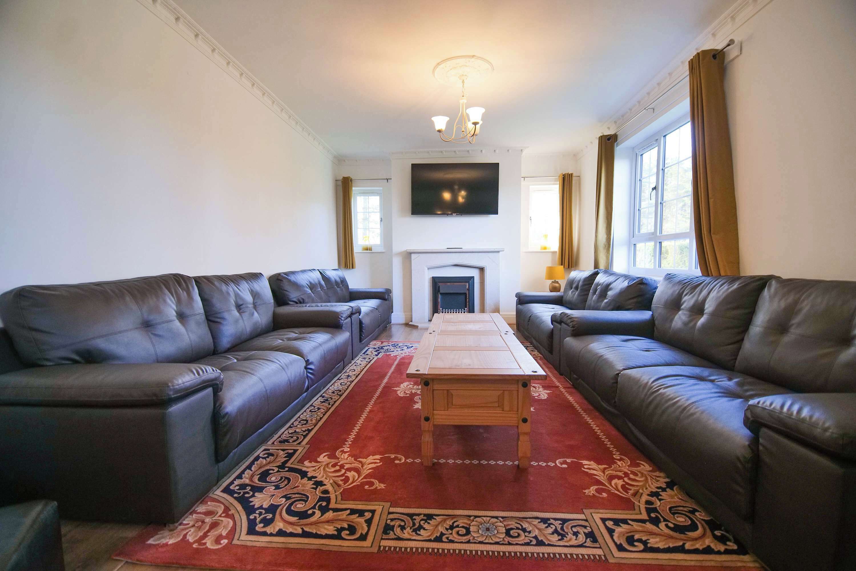A living room with two leather sofas, a wooden coffee table, a red patterned rug, a fireplace, and a television mounted on the wall.