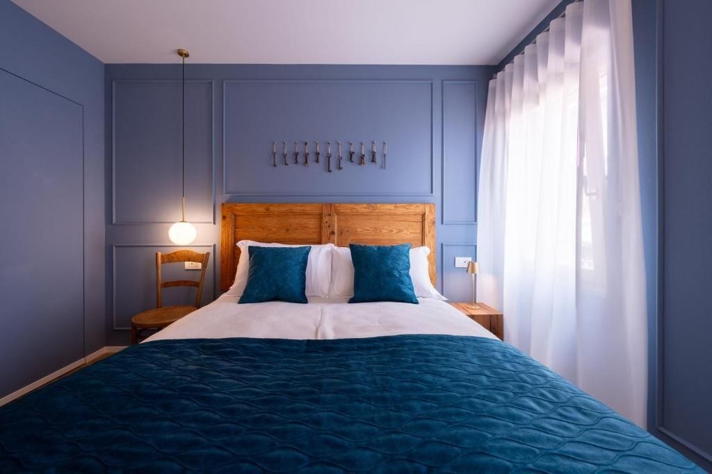 Ein Schlafzimmer mit einem hölzernen Kopfteil, einem Bett mit blauer Bettwäsche, einer Wandleuchte und einem Fenster mit durchsichtigen Vorhängen.