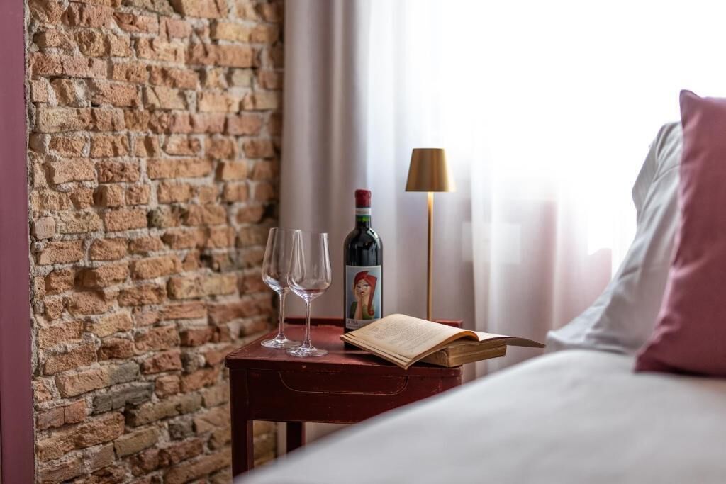 Habitación de hotel con pared de ladrillo, una mesita de noche con botella de vino y copas, una lámpara y una cama con almohadas.