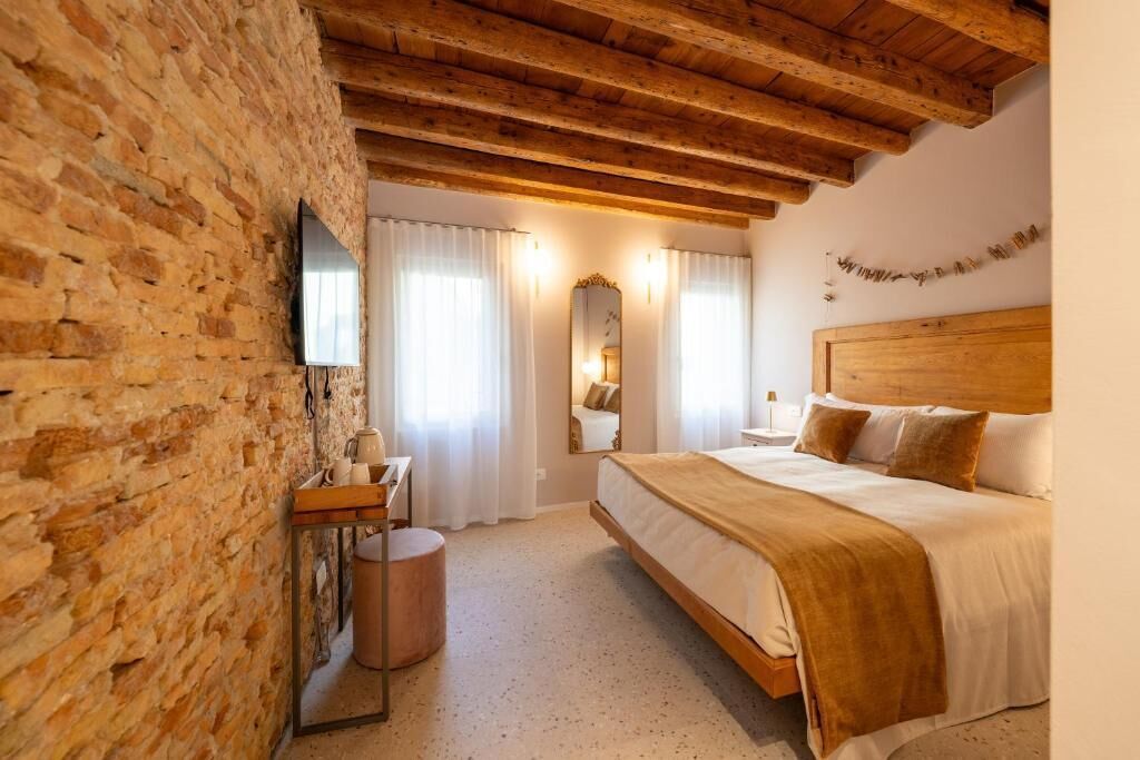 A bedroom with a wooden ceiling, a brick wall, a bed with a headboard, a bedside table, a mirror, and a television.