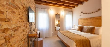 A bedroom with a wooden ceiling, a brick wall, a bed with a headboard, a bedside table, a mirror, and a television.