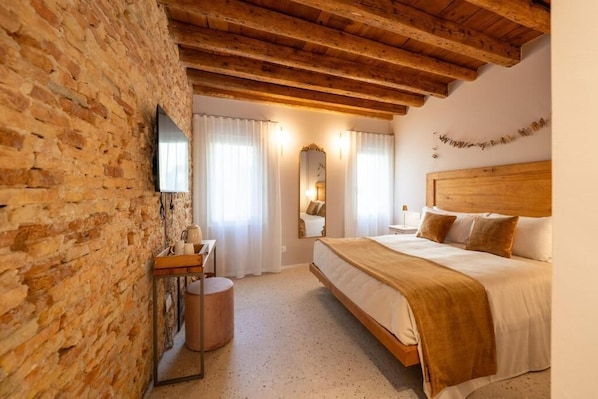 A bedroom with a wooden ceiling, a brick wall, a bed with a headboard, a bedside table, a mirror, and a television.