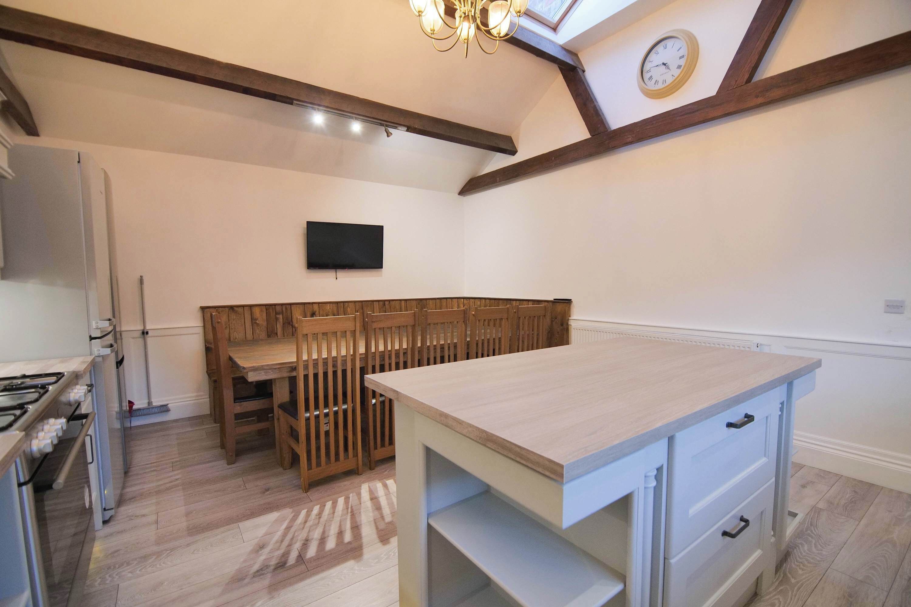 A modern kitchen with a central island, wooden chairs, a flat-screen TV, and a clock on the wall.