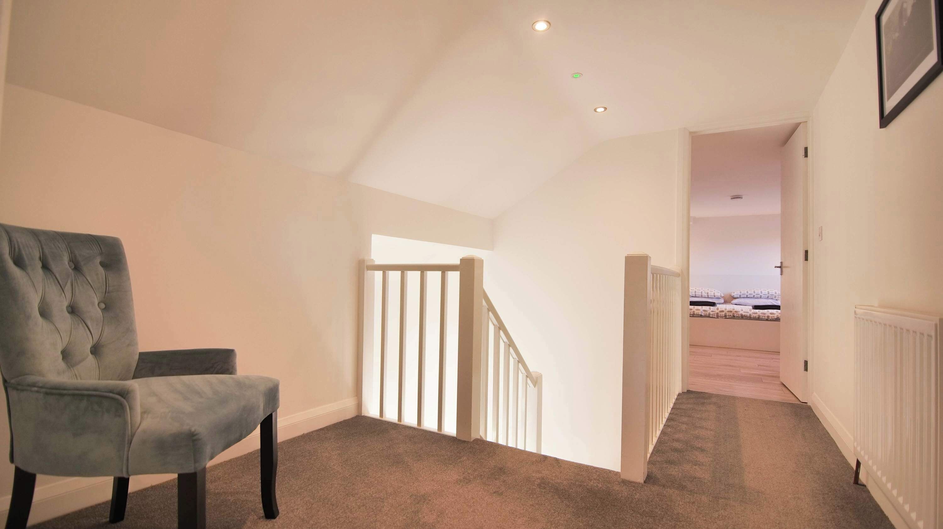 A room with a grey upholstered chair, a white staircase, and a view into another room with a bed.