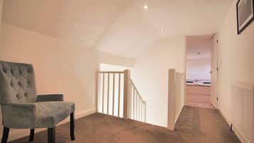 A room with a grey upholstered chair, a white staircase, and a view into another room with a bed.