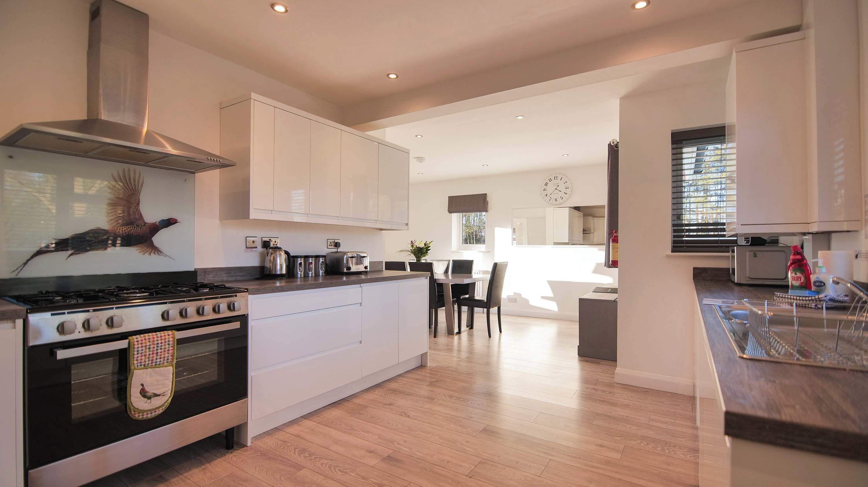 A modern kitchen with a gas stove, oven, and stainless steel range hood. There is a dining area with a table and chairs, and a large window providing natural light.