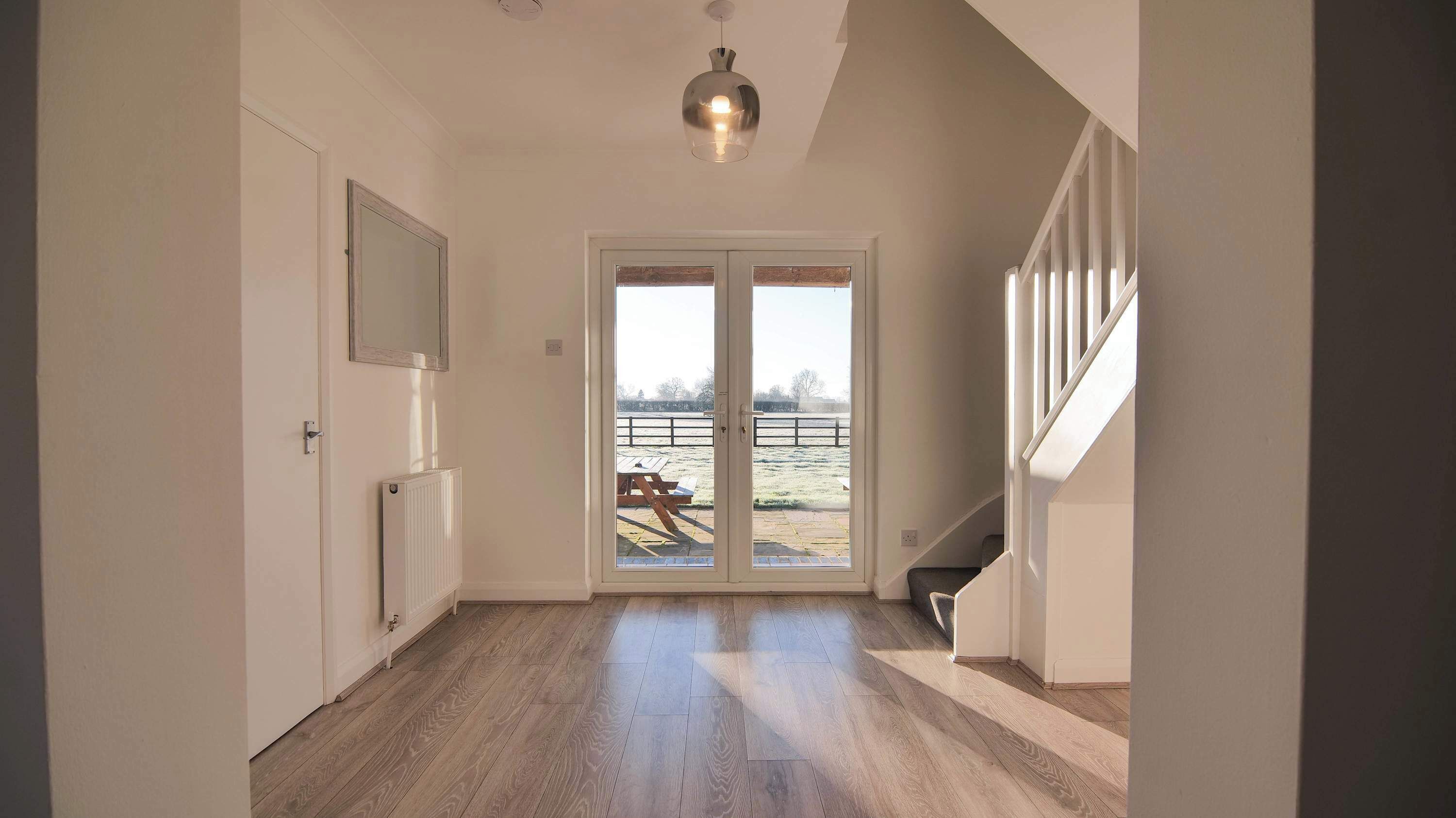 Un pasillo con piso de madera, un radiador, una puerta con espejo y vista a un área exterior cercada.