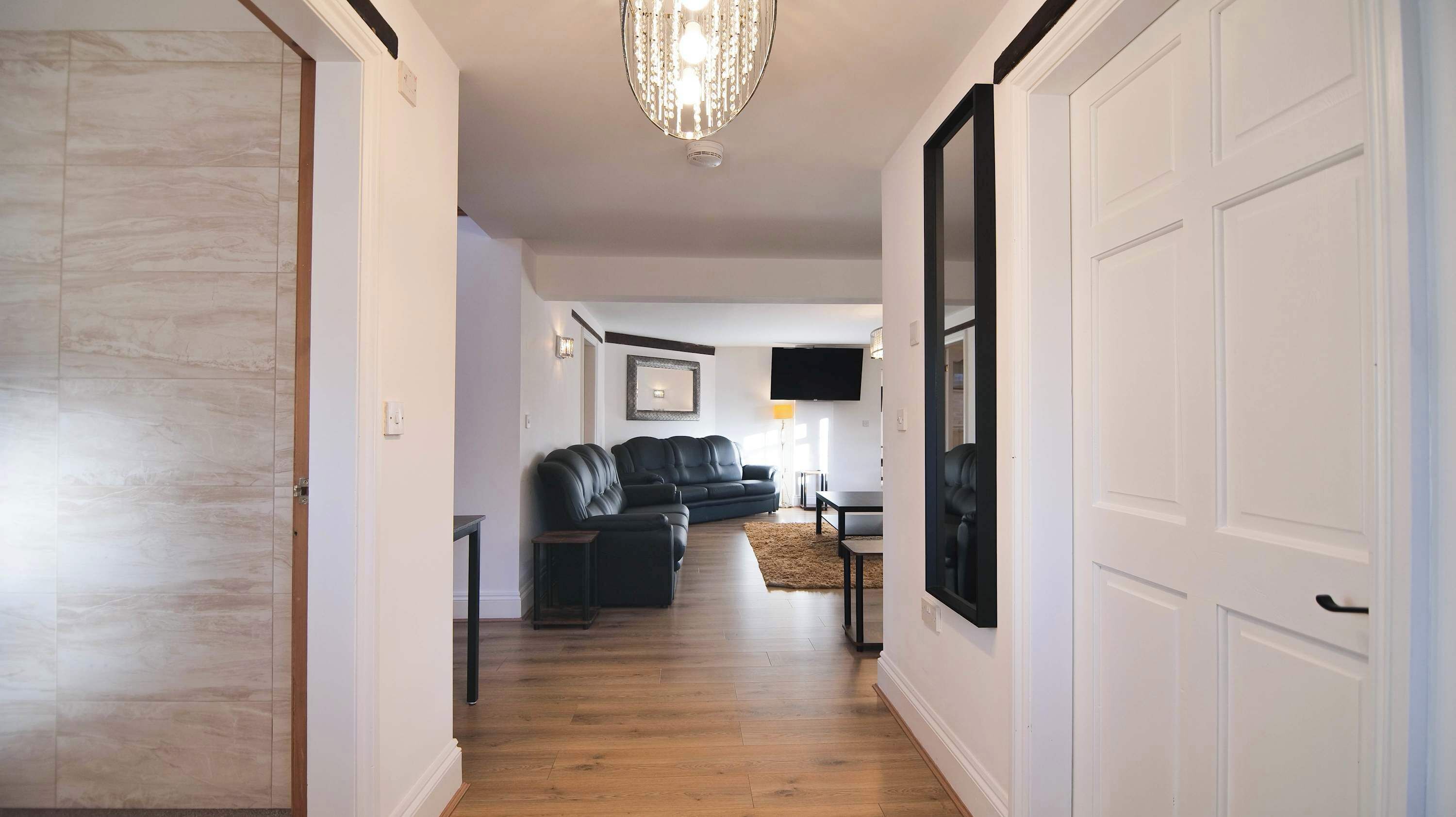 A hallway with a wooden floor, a black leather sofa, a flat-screen TV mounted on the wall, and a chandelier.