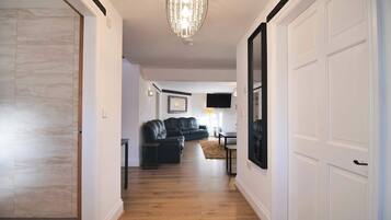 A hallway with a wooden floor, a black leather sofa, a flat-screen TV mounted on the wall, and a chandelier.