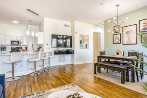 A modern kitchen with white cabinets, a central island with bar stools, a dining area with a wooden table and chairs, and artwork on the walls. - Pink Apple l Modern Comfort (Davenport)