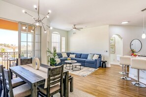 A spacious living area with a blue sofa, a dining table with chairs, a ceiling fan, and a view through large windows. - Pink Apple l Modern Comfort (Davenport)