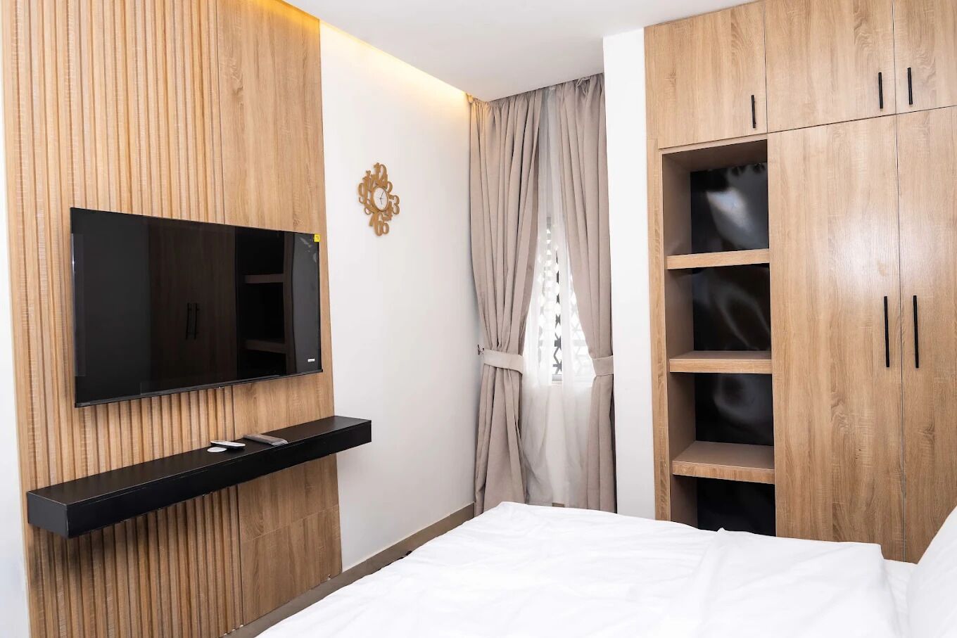 A bedroom with a TV mounted on the wall, a bed, wooden wardrobe, and a window with curtains.