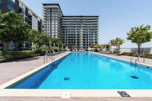 En swimmingpool med solstolar och parasoller, omgiven av träd och moderna byggnader.