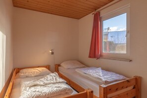 Eine kleine, einfache Unterkunft mit zwei Holzbettchen, einem Fenster mit Vorhang und einer Wandleuchte.