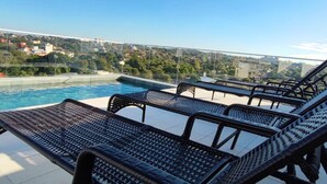 Área de piscina en la azotea con sillas de descanso y vistas a un paisaje urbano.