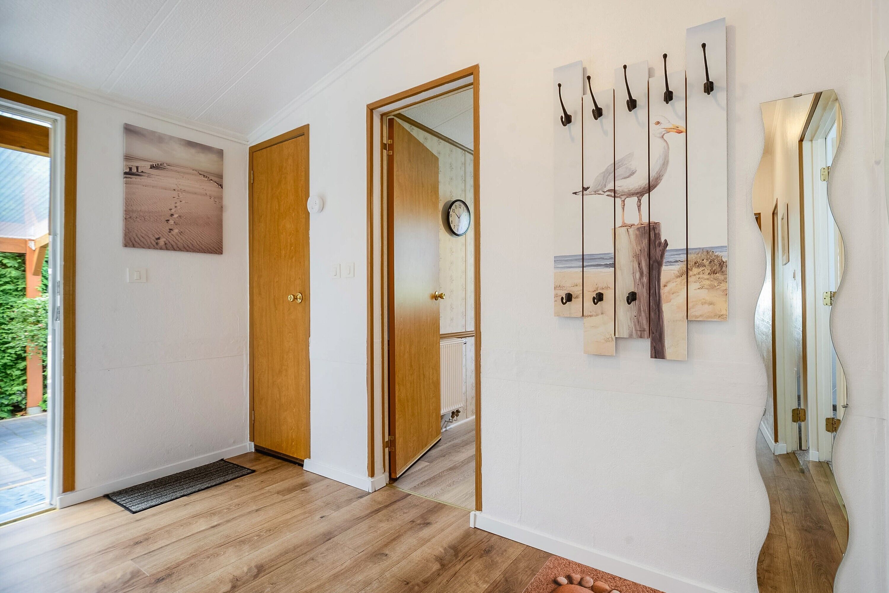 Um corredor com piso de madeira, uma porta que dá para um quarto com um relógio, e uma parede com uma pintura de gaivota e ganchos.