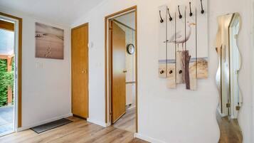 Um corredor com piso de madeira, uma porta que dá para um quarto com um relógio, e uma parede com uma pintura de gaivota e ganchos.