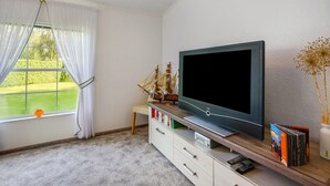 A room with a television on a wooden stand, a bookshelf with books and a CD collection, a small table with a model ship, and a window with curtains. - Rauchfangswerder Holiday Home with Private Terrace, Garden, and Wi-Fi (Berlin)