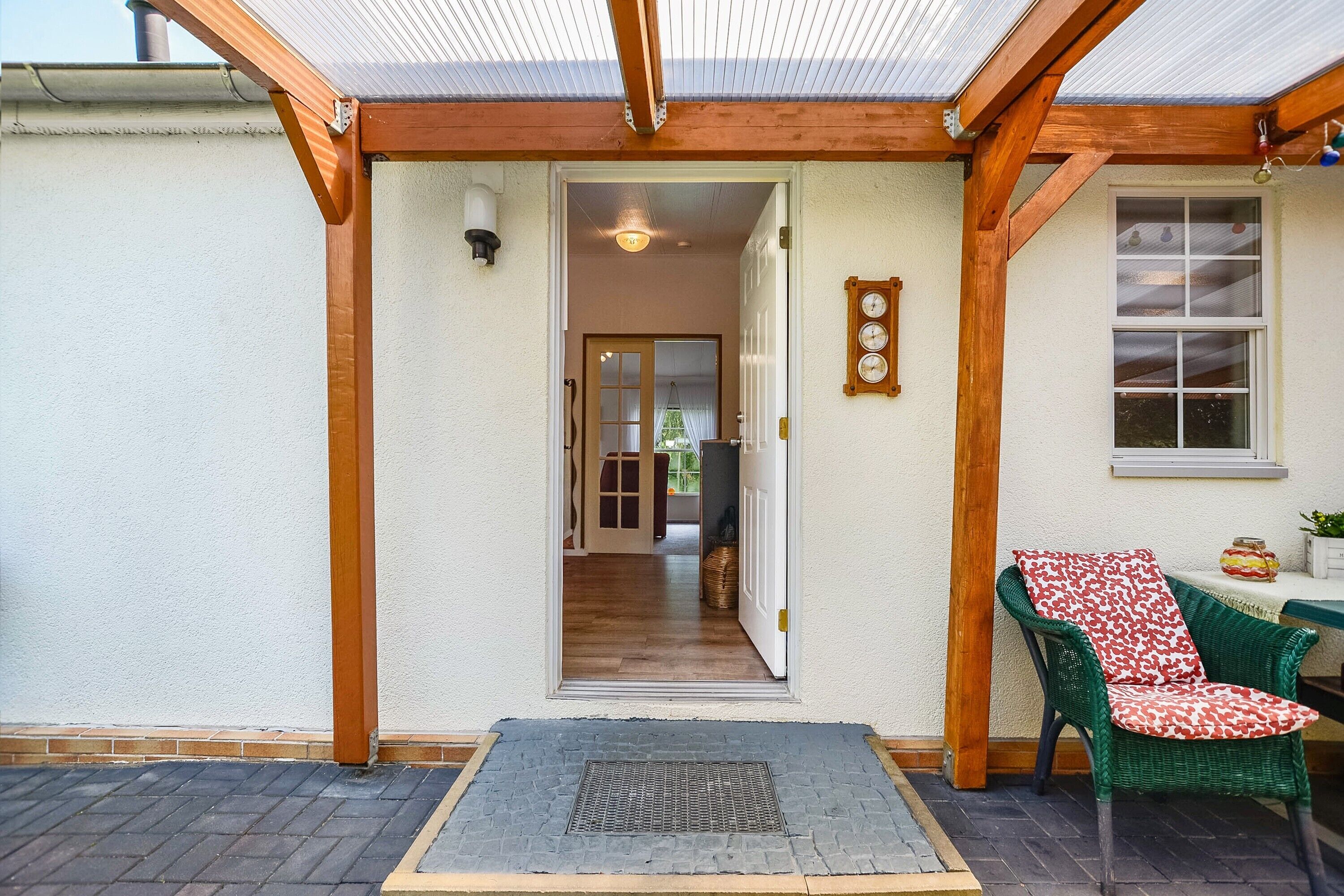 A covered entrance with a wooden frame, a door leading inside, a small table with a chair, and a wall-mounted clock.
