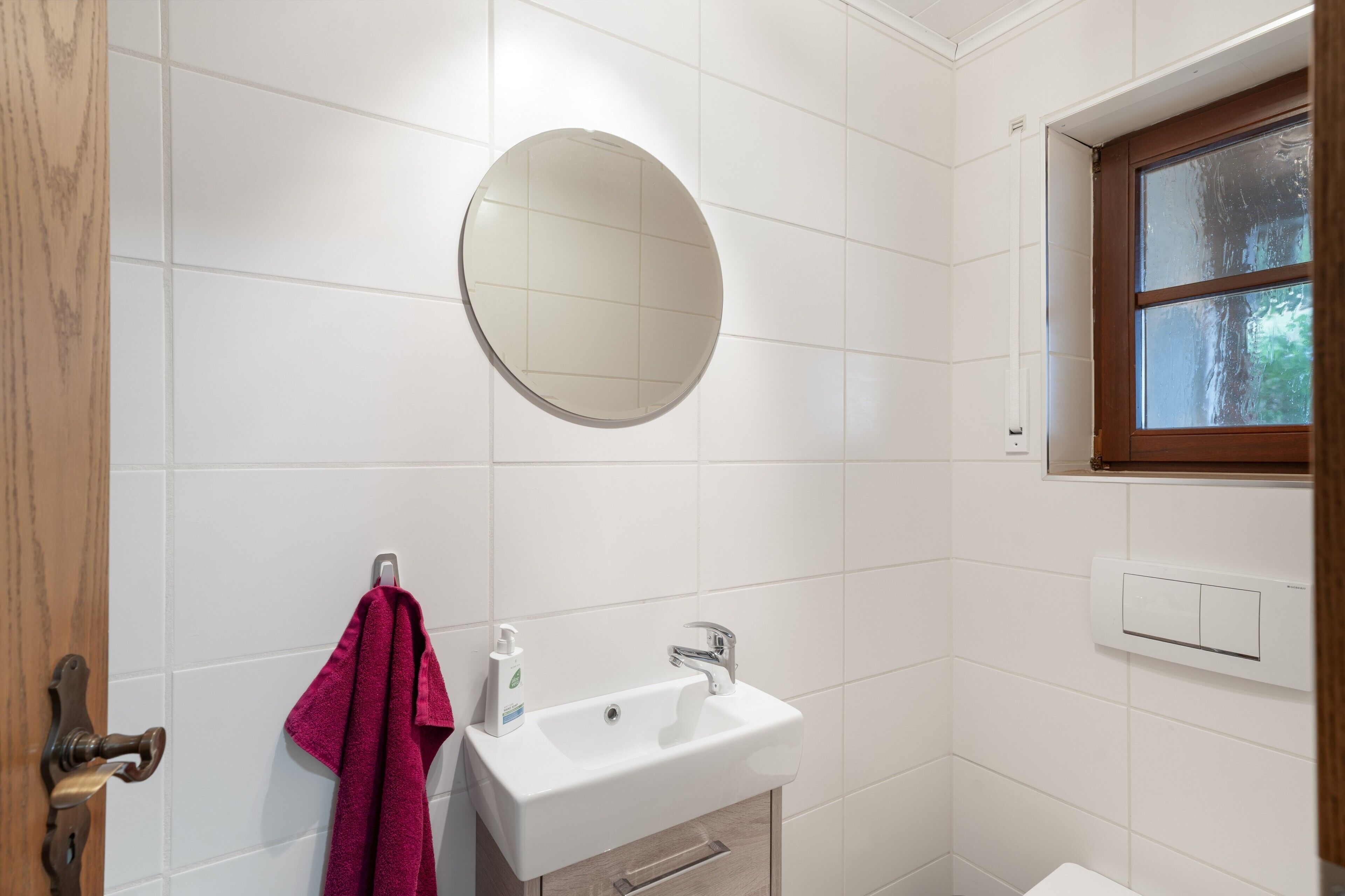 A bathroom with white tiles, a round mirror, a sink, a towel, and a window.