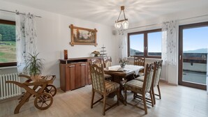 A dining area with a wooden table and chairs, a painting on the wall, and a view of the outdoors through large windows.