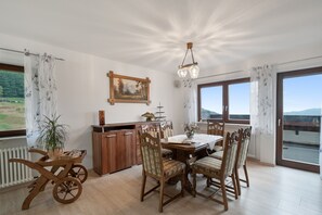 A dining area with a wooden table and chairs, a painting on the wall, and a view of the outdoors through large windows.