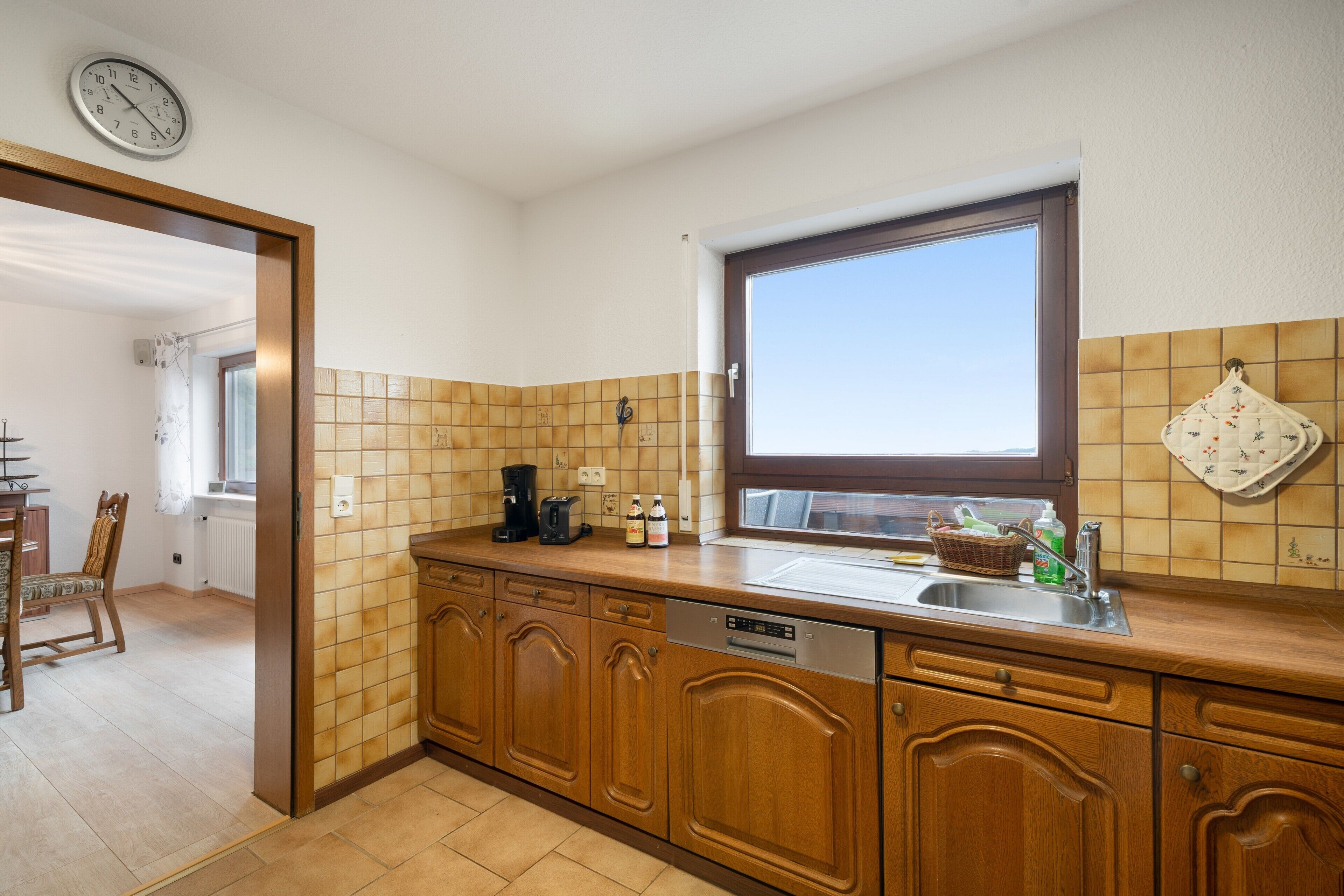 A kitchen with wooden cabinets, a built-in dishwasher, a window, and a clock on the wall.