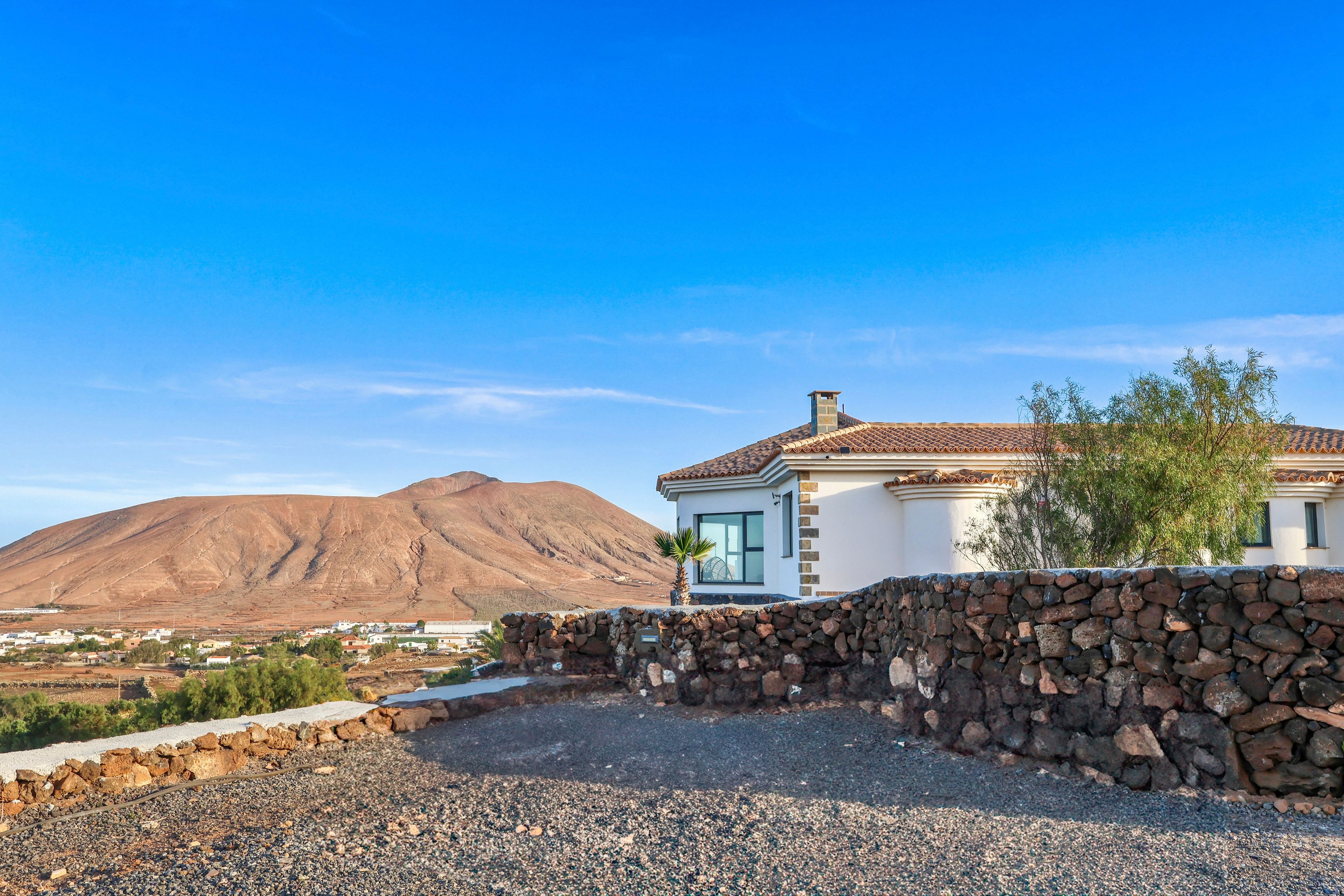 Una casa con un muro de piedra, una montaña al fondo y un cielo despejado.