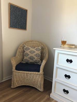 Una silla de mimbre con un cojín estampado, un vaso de líquido ámbar y un aparador blanco con tiradores negros.
