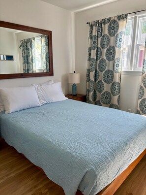 Ein Zimmer mit einem Bett, einem Nachttisch, einer Lampe, einem Spiegel und gemusterten Vorhängen.