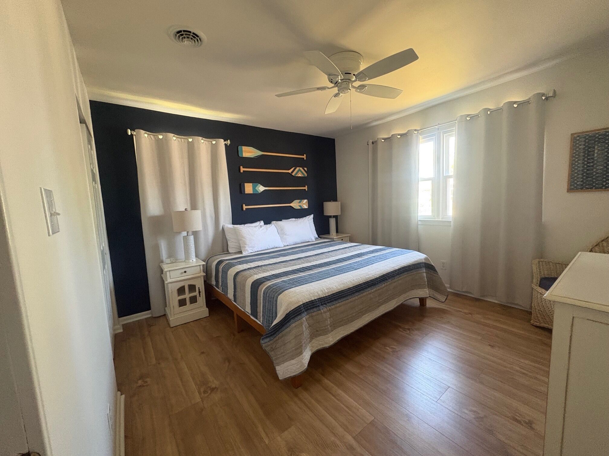 A bedroom with a bed, a ceiling fan, a window with curtains, a wall-mounted lamp, and a small bedside table.