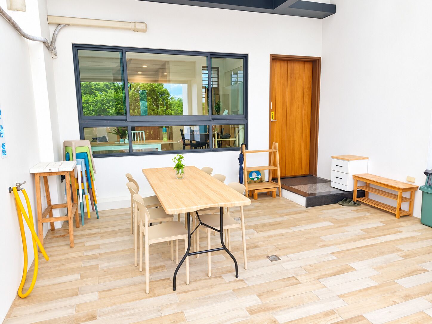 A room with a wooden table and chairs, a window with a view of greenery, a wooden door, and a small bench.