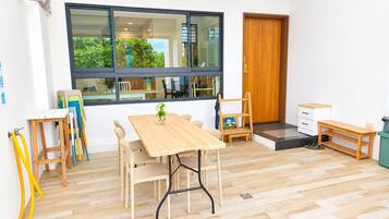 A room with a wooden table and chairs, a window with a view of greenery, a wooden door, and a small bench.