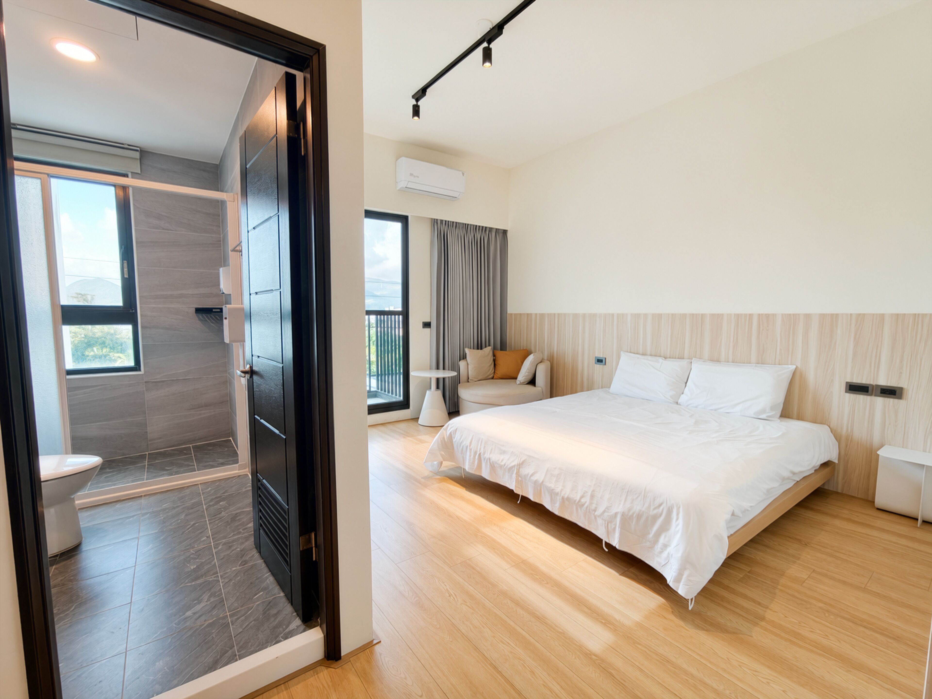 A modern hotel room with a large bed, a small seating area, a balcony, and a separate bathroom.