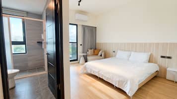 A modern hotel room with a large bed, a small seating area, a balcony, and a separate bathroom.