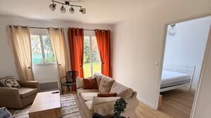 Uma sala de estar moderna com sofá, mesa de centro e uma poltrona preta. Há uma pequena mesa com uma planta e um abajur. O quarto tem piso de madeira e janelas amplas com cortinas.