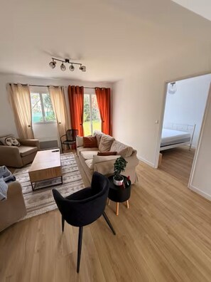 Ein modernes Wohnzimmer mit einer Couch, einem Couchtisch und einem schwarzen Sessel. Ein kleiner Tisch mit Pflanze und Lampe steht daneben. Der Raum hat Holzböden und große Fenster mit Vorhängen.