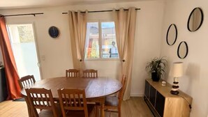 A dining area with a round table, wooden chairs, a window with curtains, a wall clock, and a cabinet with a lamp.