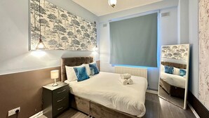 Un dormitorio con cama, mesita de noche, lámpara, radiador, ventana y espejo fijado a la pared.