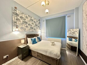 A bedroom with a bed, bedside table, lamp, radiator, window, and a wall-mounted mirror. - The London Stage Door (London)