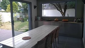 A dining area with a table and chairs, a window with a view of greenery, and a shelf with books and decorative items. - A Little Paradise with a Pool in the Heart of Nantes (Nantes)