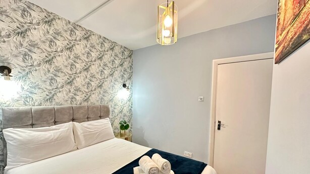 Una cama bien hecha con cabecera, una pared con diseño de hojas, una lámpara colgante y una puerta.