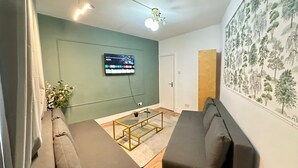 Un salón con sofá, una mesa de centro, un televisor montado en la pared y un llamativo papel tapiz con motivos botánicos.
