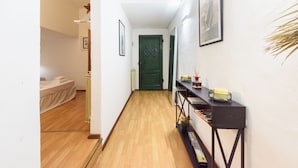 Un pasillo con piso de madera, una puerta verde, una cama con cubrecama blanco y una consola negra con libros y una vela.