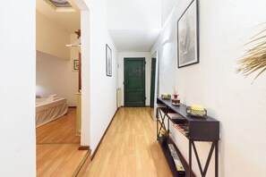 Un pasillo con piso de madera, una puerta verde, una cama con cubrecama blanco y una consola negra con libros y una vela.