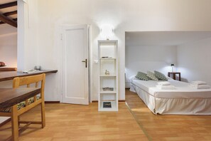 Una habitación con piso de madera, una cama con ropa blanca, una mesita de noche con lámpara, una estantería con libros y una puerta que da a otra habitación.