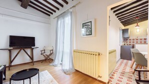 Una habitación con televisión sobre un mueble bajo, una mesita, una silla, un radiador y una alfombra estampada.